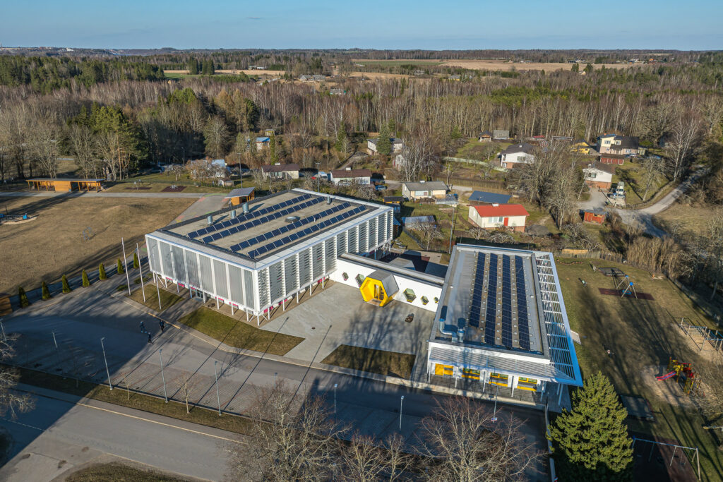 Ramsi lasteaia Taruke moodne hoone droonifotol, päikesepaneelidega kaetud katused, kollane peasissepääs ning ümbritsev mänguala ja eramud roheluses.